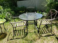 Photo showing round glass top table with grey/black metal frame and three brown metal chairs on grass outdoors, one chair visible with broken strap on seat.
