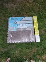 Pack of four brown Best-Step antifatigue mat squares wrapped in plastic packaging with label, accompanied by packaged white mini blinds, placed on grass