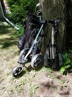 Dark colored Fairway golf bag loaded with right-handed clubs next to two collapsible golf push carts with three wheels each positioned outdoor near tree base