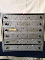 Front view of metal cabinet with five drawers closed, textured galvanized finish, black knobs on each drawer, adhesive label remnants on front.