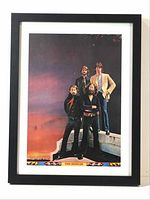 Framed vintage print showing the Beatles band members standing with arms crossed and in casual poses against a colorful sky background.