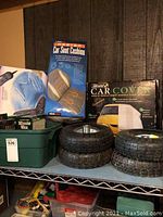 Shelf with ObusForme heated car seat cushion box, Sidewinder car cover box, four trailer tires stacked in two sets including rims, and various Turtle Wax car care products in green plastic bins.