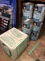 Stack of six 1-gallon paint cans and one more paint can beside them on the wooden floor with a white box near by