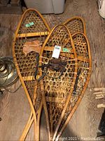 Two pairs of wooden snowshoes side by side on wooden floor, showing overall frames and woven decking with leather harnesses attached.