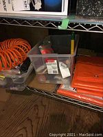 Wide view showing orange tarps in original packaging and folded tarps on shelf, plastic bins with paint supplies and hardware, and a garden hoe handle visible in hardware bin.
