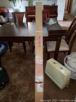 Photo showing the original packaging box of Elite Screen projection screen sitting on a carpet next to furniture. The long box is labeled 'Projection Screen' with visible tape and wear marks.