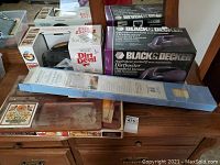 Photo showing two handheld vacuum boxes (Black & Decker Dirtbuster and Dirt Devil Scorpion), long package containing mini blind, wooden tray with decorative design on table, and Cornell hot electric tray.