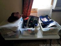 Three clear plastic clothing bins on table, one with black shoes and socks, one with folded pants including blue denim with tags, and one with assorted clothing including men's and ladies' items.