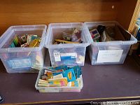 Four clear plastic storage bins filled with an assortment of office supplies including pens, pencils, markers, binder clips, and stationery items.