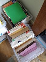 Two clear bins filled with various office supplies including file folders and paper, viewed from above at an angle.