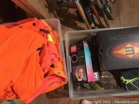 Two bins with orange safety jackets, camo clothes, and various gear including a box and bags above fishing rods.