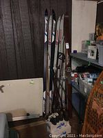 Multiple vintage cross country skis standing upright including Ross Country, Germina, and Medalist brands with poles and leather boots at the base, shown against a wall and shelves with miscellaneous items.