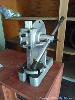 Side view of the gray cast metal jewellers press showing the vertical ram, hand lever, and rotating base.