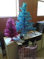 Photo showing two metallic artificial holiday trees, a tall blue tree and a shorter dark pink tree sitting on a wooden surface alongside assorted holiday decorations and wrapping.