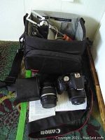 Black Canon camera bag open with white cables, manuals, red-handled tool, camera body and lens on patterned carpet.
