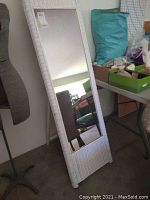 Full view of the tall white wicker cheval mirror standing upright reflecting surroundings.