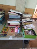Table showing multiple stacks of folded vintage fabrics in various colors and patterns, green boxes with lace and ribbons, vintage sewing patterns, a few cones of threads and small scissors.