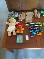 Overview photo showing Cabbage Patch doll, Fozzy Bear plush, Thomas & Friends books, train cars, track pieces, and ME Reader toy arranged on a wooden table.