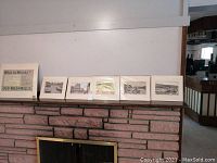Photo showing six matted prints lined up on a brick fireplace mantle, each protected with plastic.