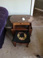 Foot stool covered with black cloth featuring a floral needlepoint wreath design. Curved wooden legs. Worn condition.