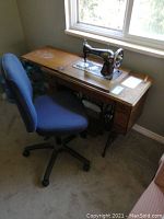 Wide view showing vintage Singer treadle sewing machine in wood cabinet with blue office chair adjacent.