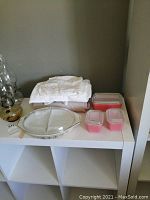 Three covered pink Pyrex containers, vintage linens, and a Pyrex serving dish displayed on a white shelf.
