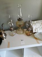 Three glass hurricane style oil lamps arranged on a table with some additional items nearby. Two lamps have clear glass and rounded bases; one lamp has a yellowish hexagonal textured base and is taller.