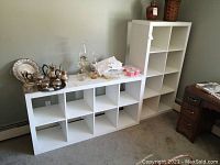 Two white shelving units against wall, one tall with 8 cubicles and one horizontal with 8 cubicles, various decorative and household items on top and inside.