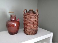 Two vintage ceramic jugs displayed on a white shelf. One jug is glossy brown glazed with a wide body and metal handle. The other jug is enclosed in a woven wicker basket with looped handles.
