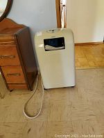 Full view of Danby portable air conditioner on wooden floor beside wooden dresser, showing unit on wheels and attached power cord.