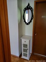 Photo of the white cabinet with drawer and glass-paneled door with shelves inside, placed beside a door.