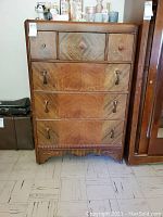 Front view of six-drawer wooden dresser showing three upper drawers with round Bakelite knobs with metal center accents, and three lower drawers with Bakelite handles. Wood veneer has a light-medium brown color with a pattern. Carved decorative wood trim along bottom apron.