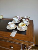 Six bone china teacups each paired with a matching saucer, displayed on a wooden surface with varied floral and gold designs.