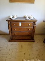 Front view of solid wood filing cabinet with three drawers and decorative carved columns on sides. Surface holds several teacups and saucers. Cabinet shows scratches and wear.