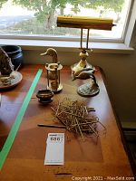 Brass desk lamp with dolphin base, Ronson Crown lighter, brass goose bust, brass candle holder, small brass container, and assorted brass hooks on wooden surface near window.