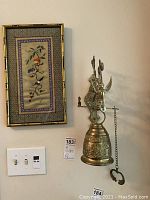 Framed embroidery artwork hanging beside a brass wall-mounted bell with decorative bracket; view includes switch plate under items.