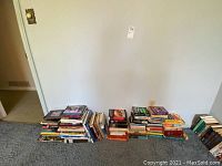 Wide view of assorted books stacked in three piles on carpet against a wall, showing the quantity and condition of the lot.