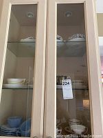 Two glass shelves inside wood cabinet showing multiple sets of vintage teacups and matching saucers including blue Fire King pieces on the bottom shelf