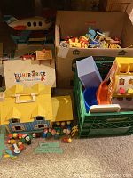 Wide view of Fisher Price cash register, airport toy, Play Family School house, blue/yellow playhouse, and wooden peg figures spread around.