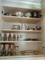 Cupboard shelves with multiple silver plated bowls, pewter beer mug, gravy boat, and trays.