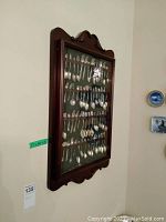 Side view of the wooden display case containing multiple collector spoons hung vertically on two horizontal bars inside, wall mounted.