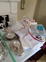 Photo showing neatly folded white linen napkins and tablecloths stacked on the table alongside shiny silver plate trays and bowls. Fabric oven mitts with floral embroidery and a red-edged round bowl with some small items inside are visible.