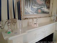 Silver plate three-arm candelabra with blue candles and two glass candle holders with blue candles on a white mantel under a framed painting.