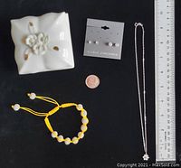Photo showing small white ceramic trinket box with floral design, sterling silver chain necklace, cubic zirconia stud earrings card, yellow fiber bracelet with cubic zirconia beads, and a coin for scale with ruler.