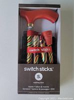 Front view of folding walking stick in original box showing red wood handle and carnival pattern on folded aluminum shaft with label 'switch sticks'