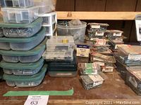 Multiple stacks of green and clear plastic storage containers filled with assorted nails and screws, small clear plastic boxes, and other packaging on a wooden surface.