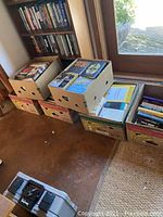 Six boxes filled with assorted books, stacked in a room near a window.
