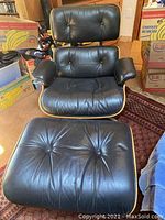 Front view of black leather Eames Lounge Chair and matching ottoman on rug amid packed boxes