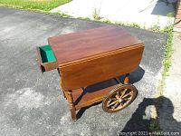 Wooden tea wagon placed outdoors showing one side drawer open and two folding side panels folded down.