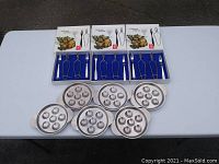 Image showing 6 stainless steel escargot dishes, 6 escargot forks, and 6 pairs of tongs laid out on a white table.